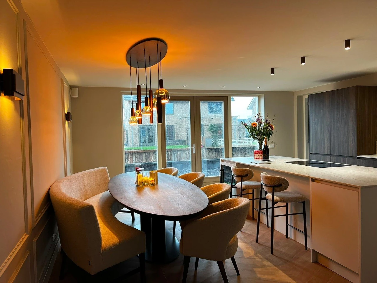 Eetkamer met houten tafel, wandverlichting en open keuken met bar